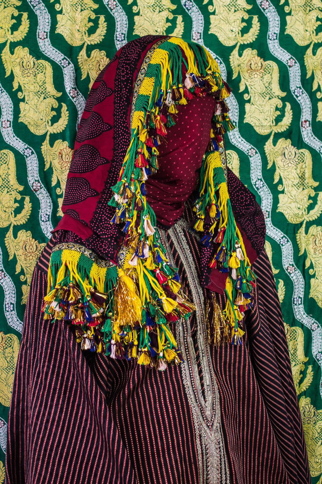 Kadia, BORDERLAND series, by Alia Ali. 2017. A model is draped in colorful fabric and tassels which completely obscures their form, photographed against a pattered fabric background