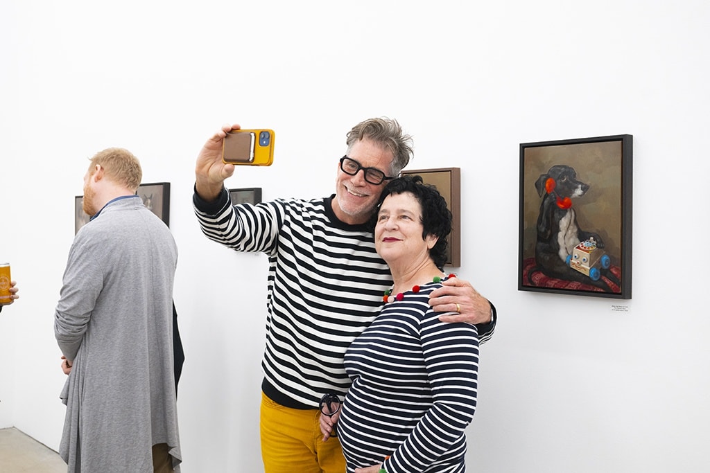 Two people in striped shirts take a selfie in front of Alison Friend's paintings in an art gallery. 