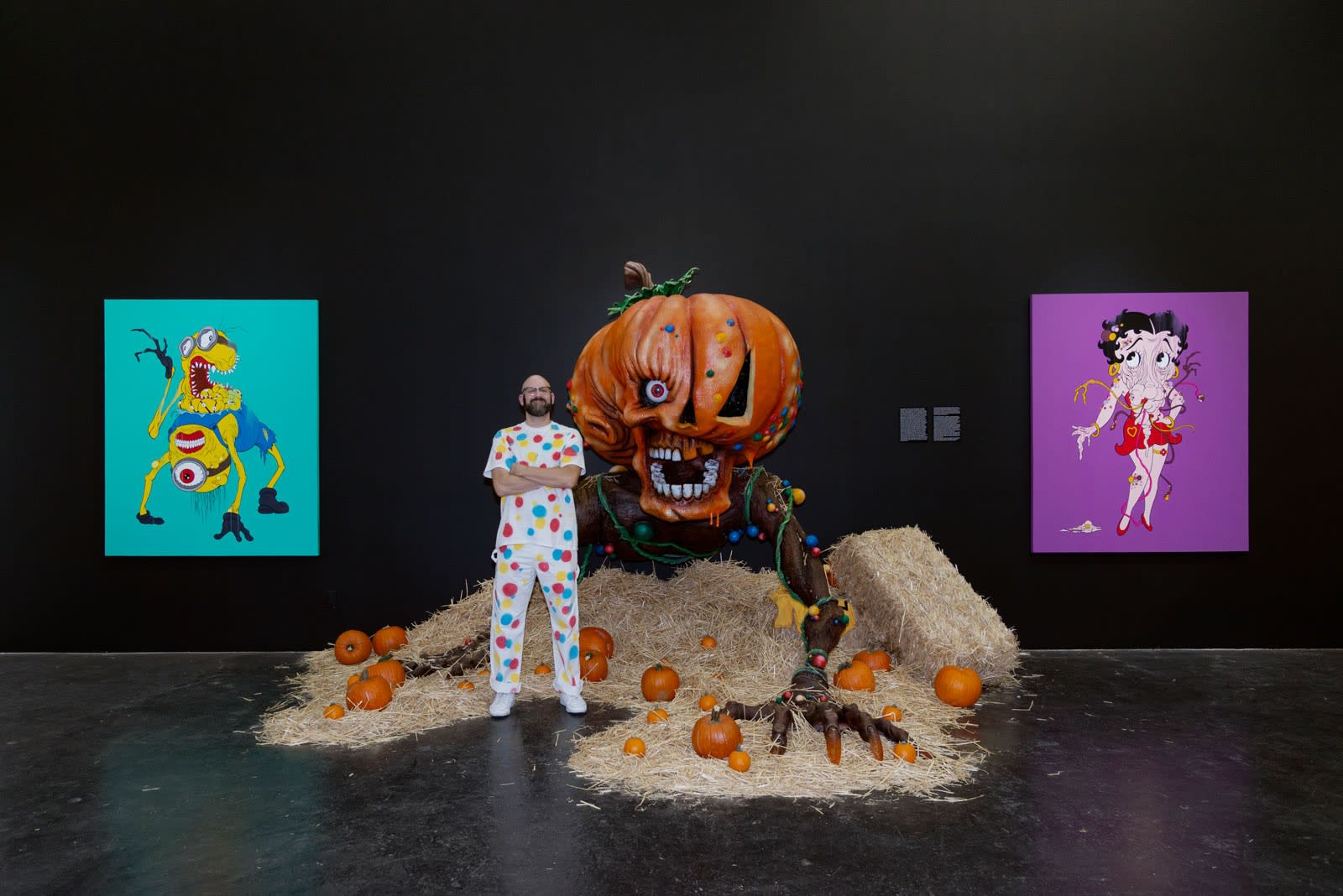 Artist Alex Pardee standing in front of Giant Pumpkin sculpture with straw and pumpkins on ground and two large paintings on the wall in background