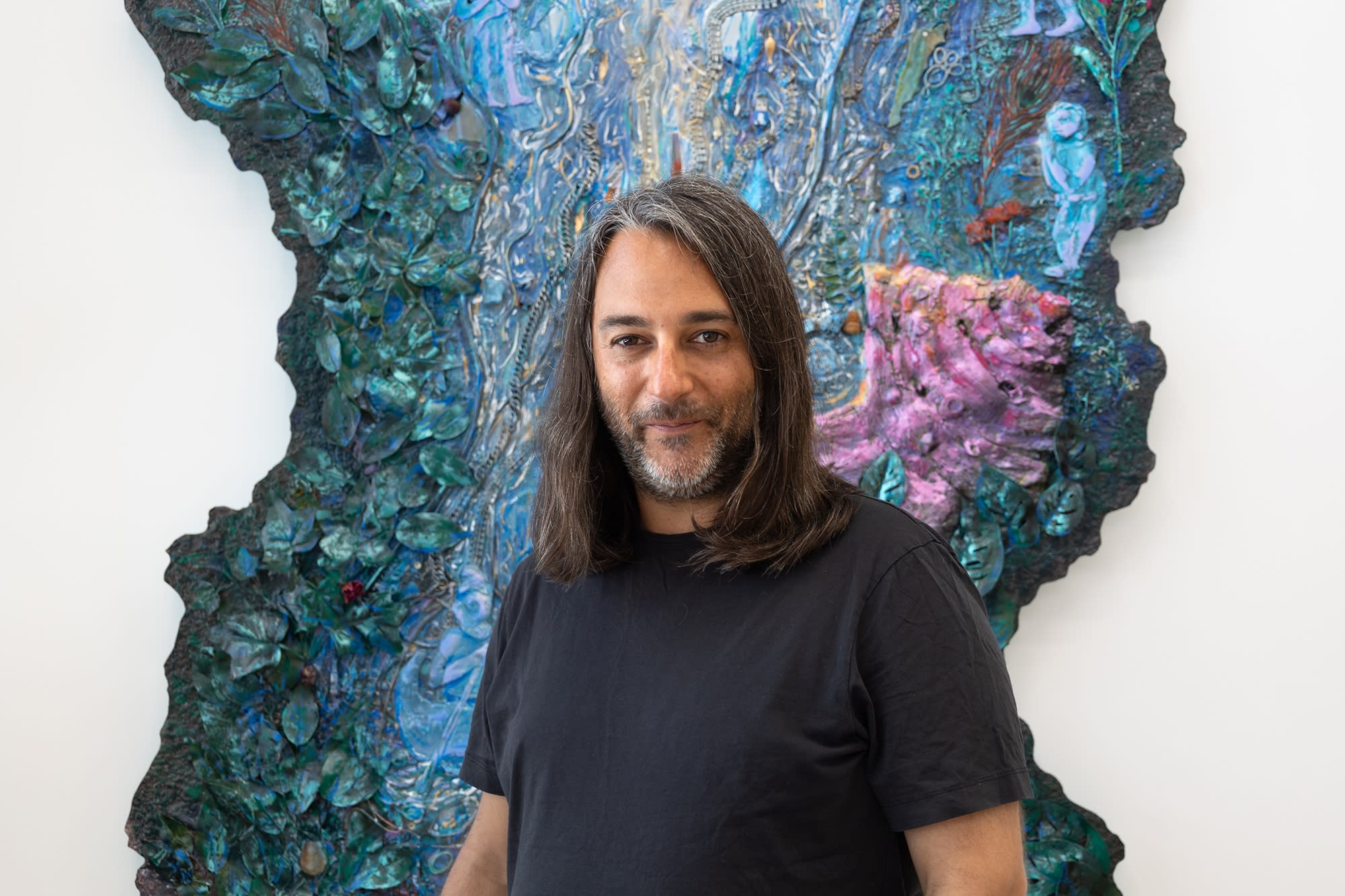 A man with olive skin and dark brown hair to his shoulders wears a silly smirk while standing in front of a blue sculptural artwork on a white wall.