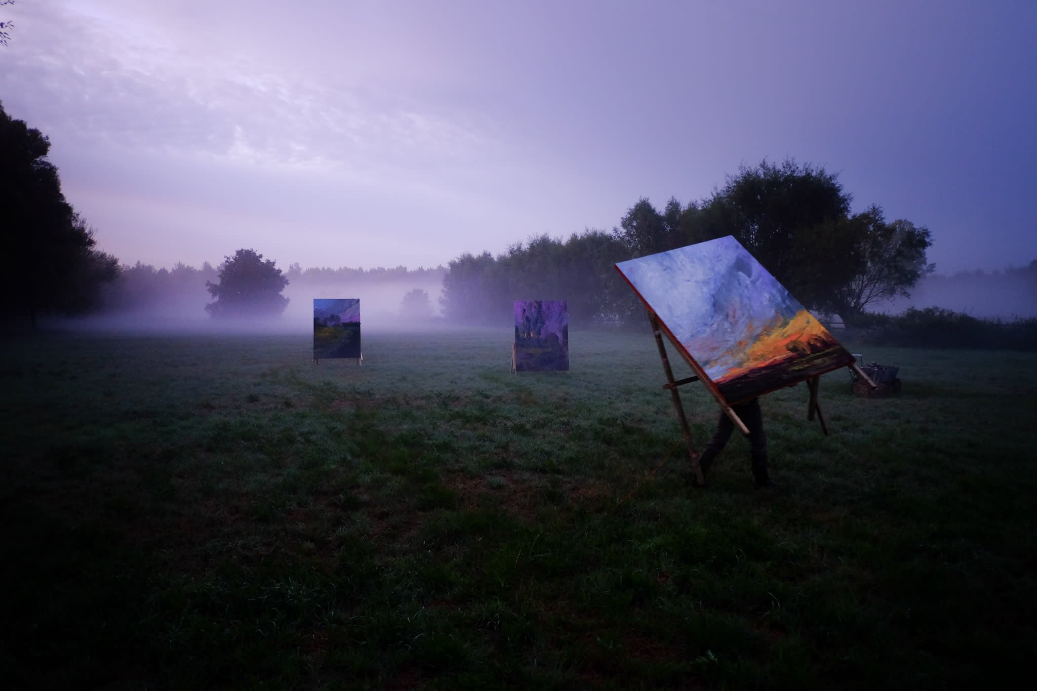 Large-scale paintings by Nils Verkaeren standing in a misty field at dawn. The artist is partly visible behind a canvas in the foreground, working in situ with trees and fog in the background.