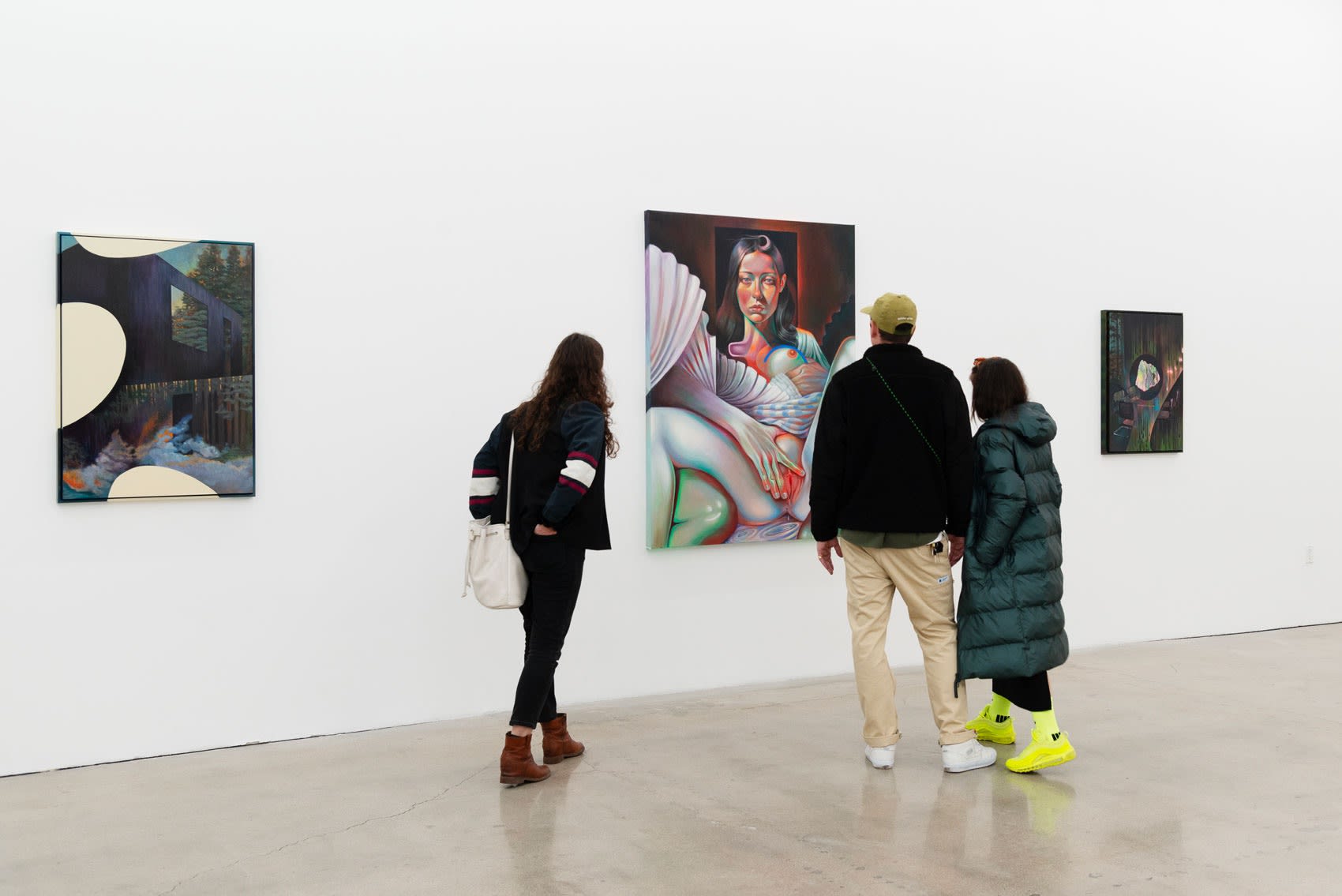Three people stand around a painting of a nude woman with her legs spread open in an art gallery. The painting, rendered in rich, psychodelic colors, is large and hangs on a white wall. 
