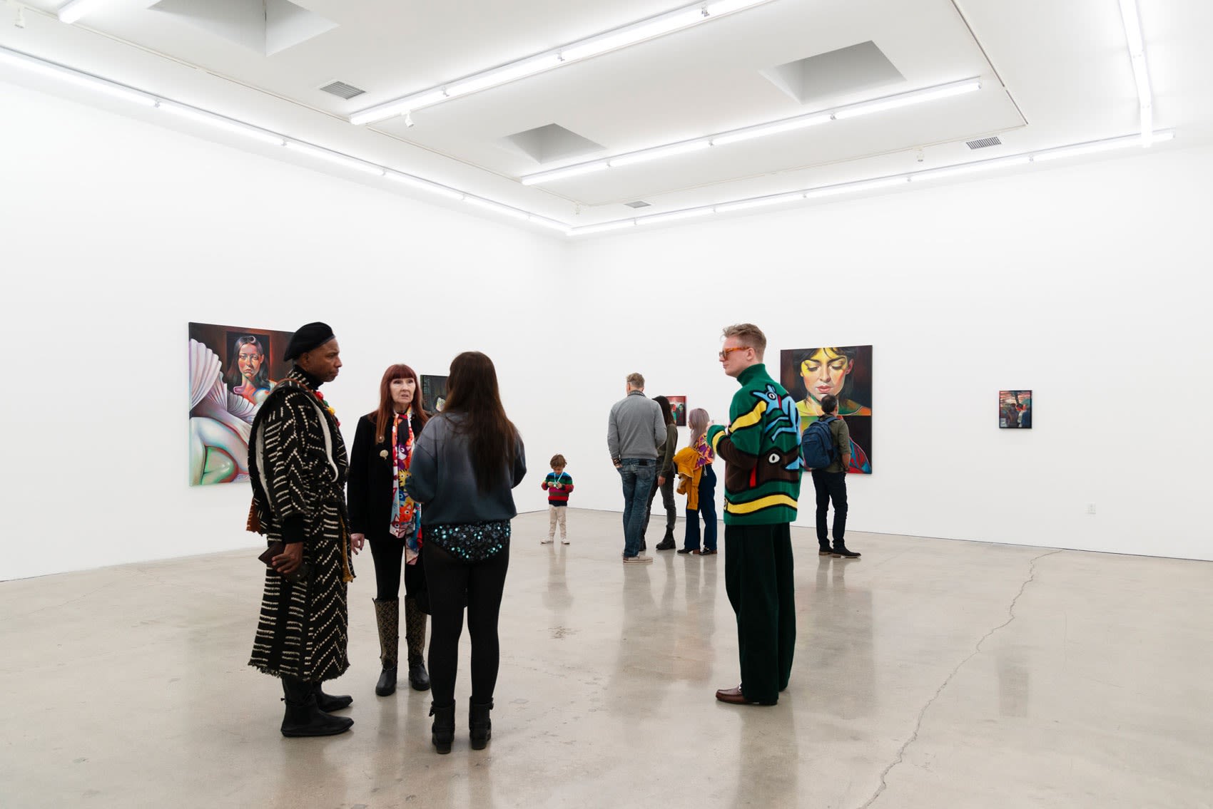 A crowd of people gather and converse inside the opening of Martine Johanna's solo exhibition A Particular Ghost. The exhibition shows paintings of women on a white wall, the gallery has some natural light and a concrete floor. 