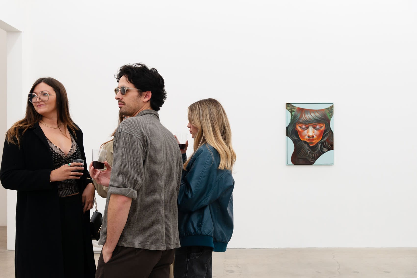 A group of young people in short sleeve shirts converse in a white wall art gallery during an opening reception. A painting of a young woman with brown hair, looking down, hangs on the white wall behind them. 
