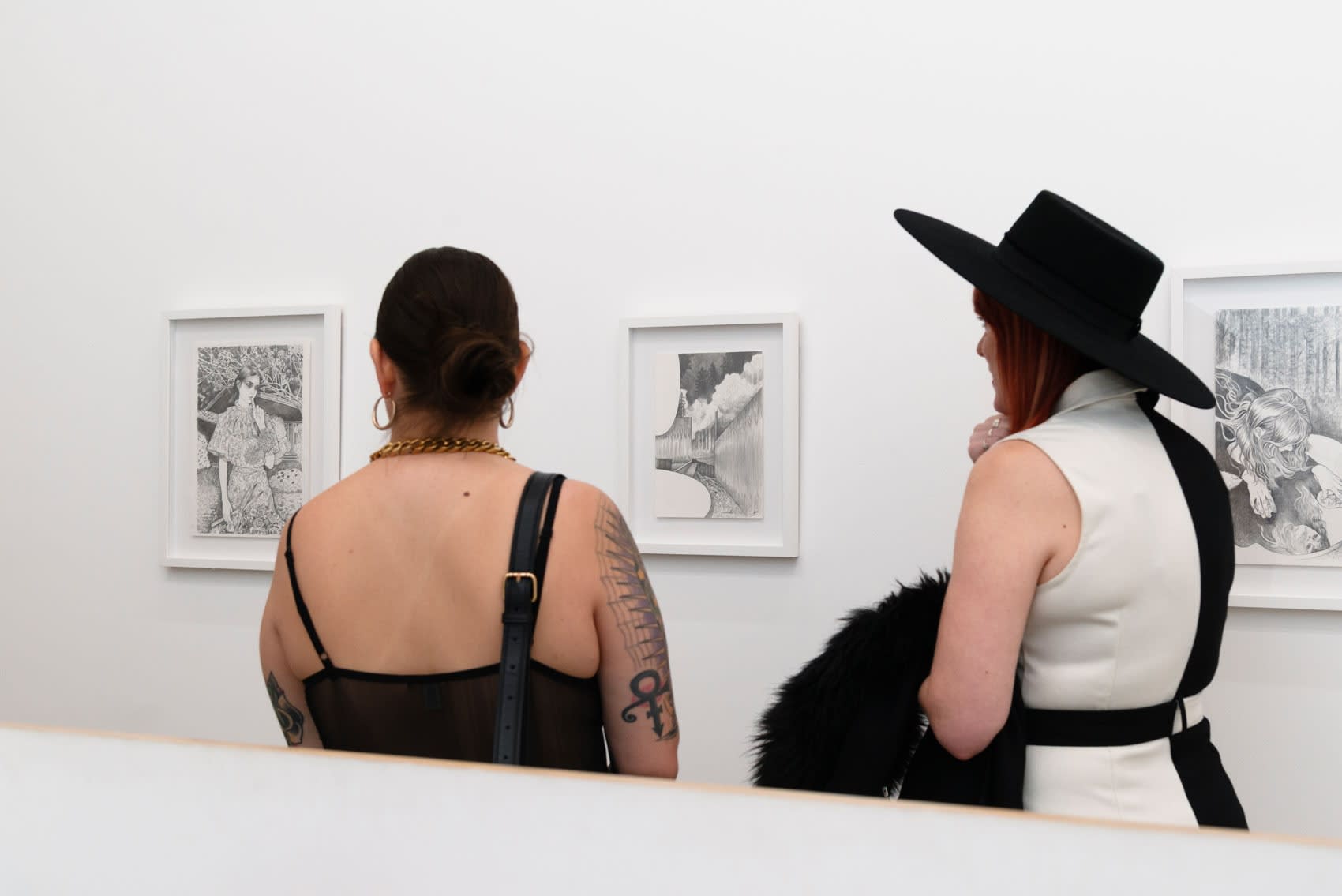 Two women stand in front of Martine Johanna's black and white drawings of young women. The drawings are framed and on a white wall in the reception area of an art gallery. 