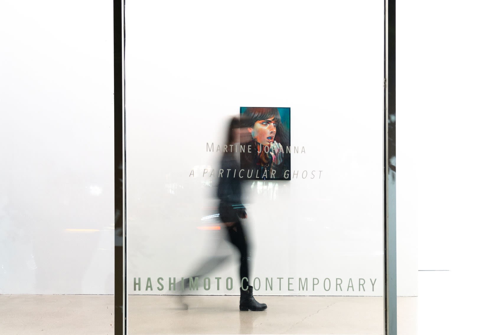 A long exposure shot of a woman walking past a painting of the head of a woman taken from outside of the art gallery. The woman walked down a hallway with floor to ceiling windows, passing the text 