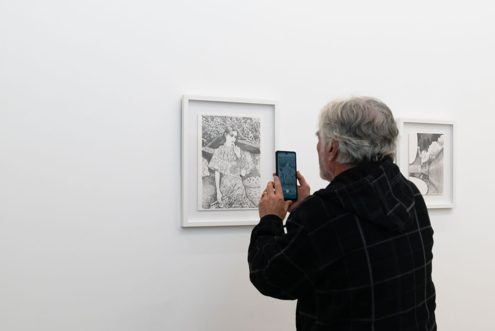 A man takes a photo of a black and white drawing on a white wall. The drawings are by the Dutch artist Martine Johanna, and depict young women doing various stereotypical activities (gardening, crying, etc.)