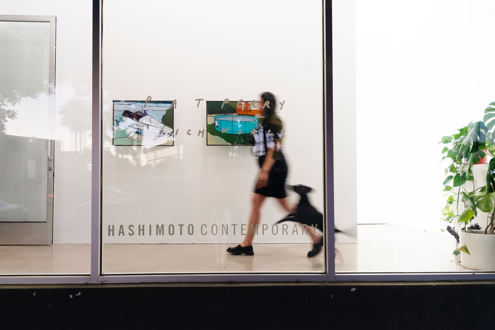 A long exposure shot from outside the gallery shows blurry people walking past two stained glass artworks on the wall, behind the art gallery's window, which reads 