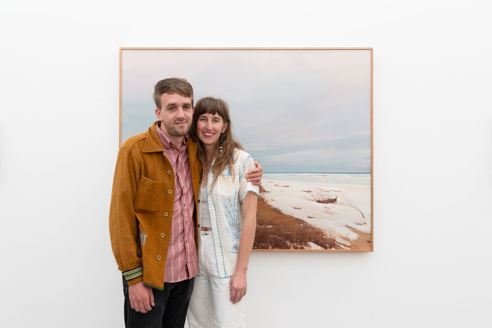 Pat Perry and his partner Rosemary Brown hug in front of one of Pat's paintings inside a large art gallery with white walls and concrete floors. Pat is a dirty blond white man wearing an burnt orange sweater and Rosemary is a dirty blond white woman wearing a white shirt with blue embroidery. 