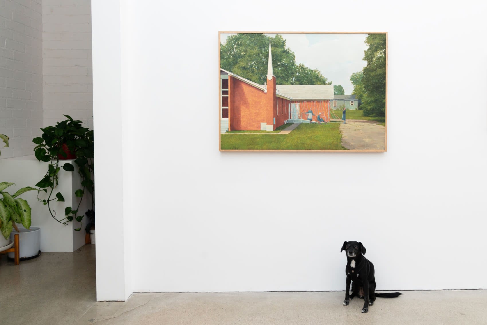 Meera, a small black dog with nervous tendencies, sits in front of Pat's painting of amish women roller blading in front of a red church.
