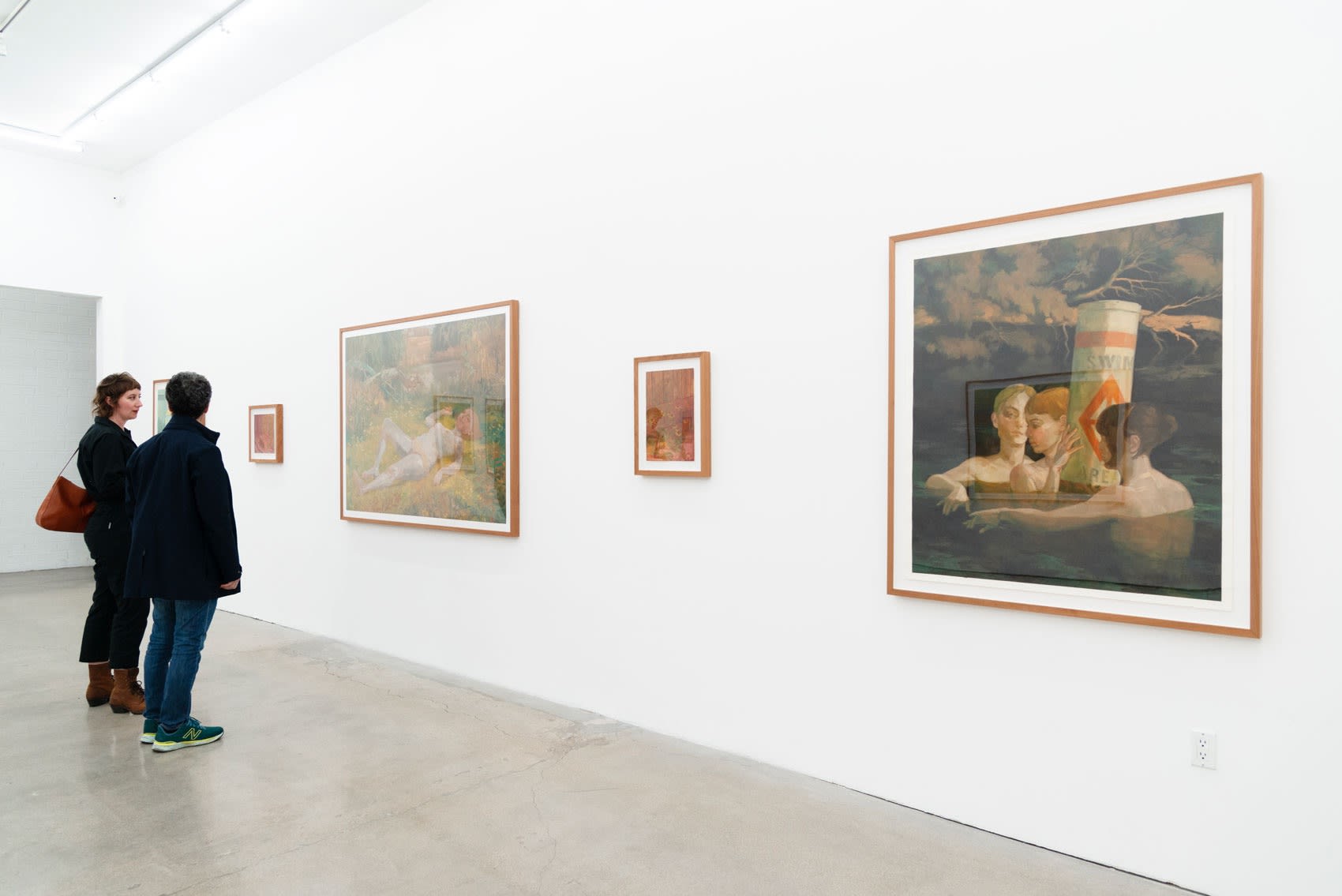 Two women stand in a white wall gallery with concrete floors looking at a clustering of framed paintings of languid girls either near a body of water or inside the dark, black water holding onto a buoy.