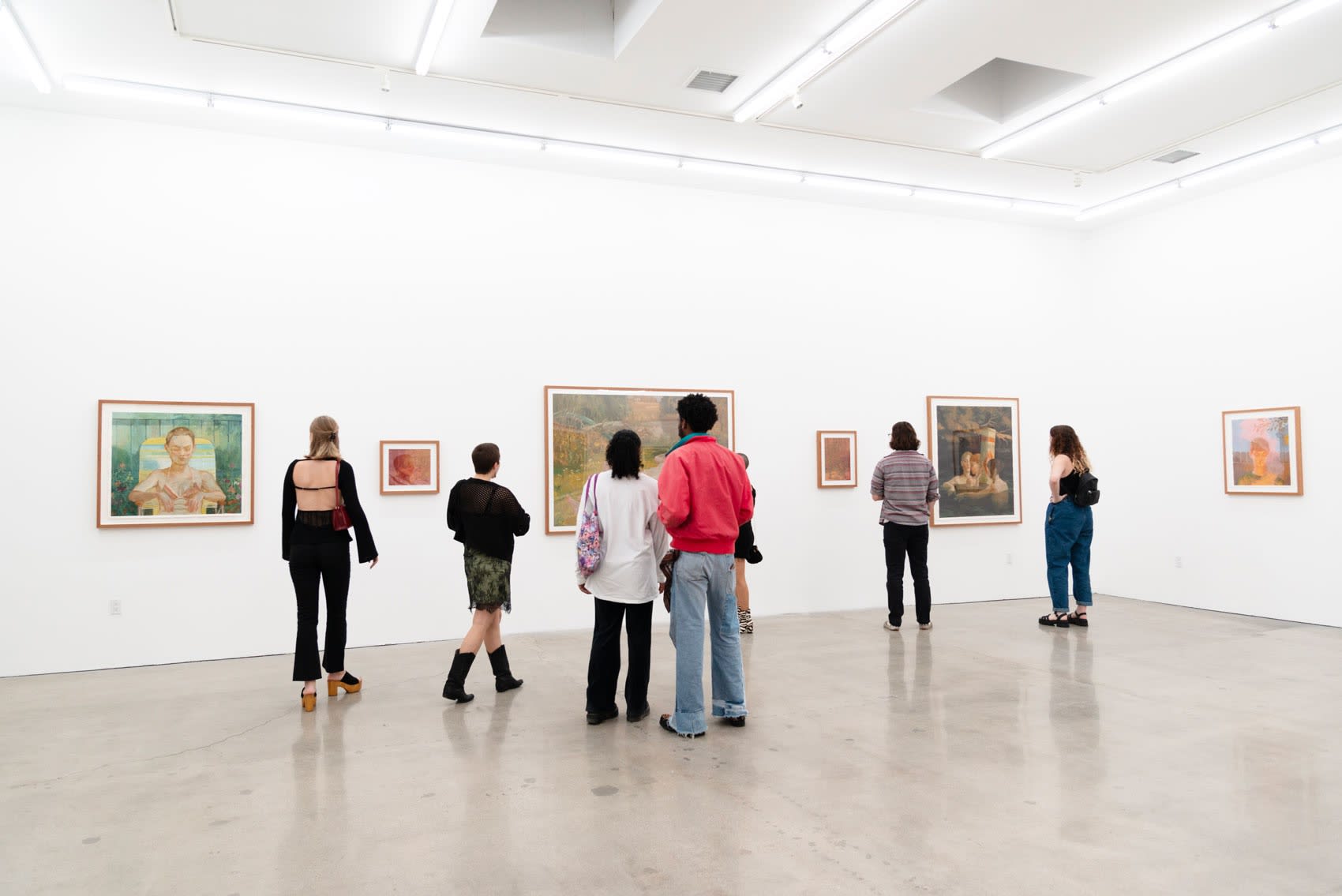 A group of young people stand in a large white wall gallery looking at Rachel Gregor's framed paintings of teenage girls going in their homes or at the park in the last days of summer before highschool. 