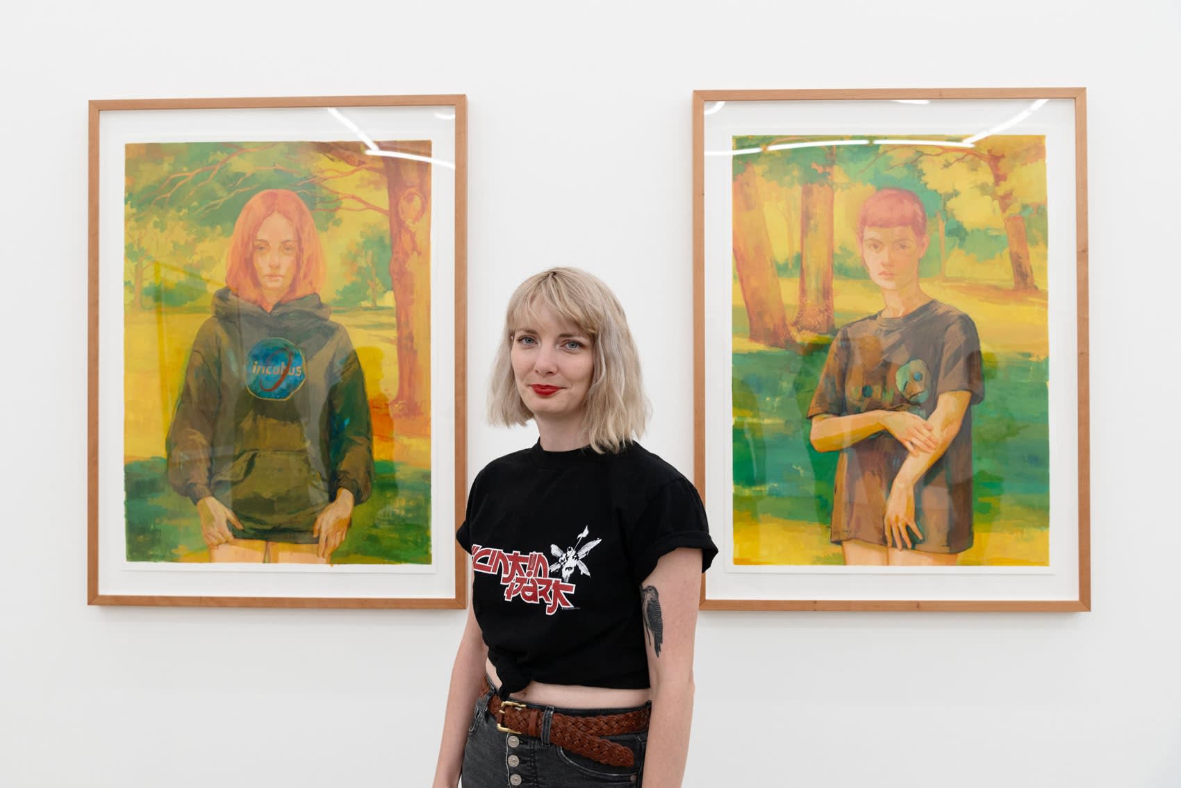Rachel Gregor, a white woman with bleach blond hair, stands in a white wall gallery between her two paintings of teenage girls wearing Nu Metal band hoodies outside on a late summer day. 