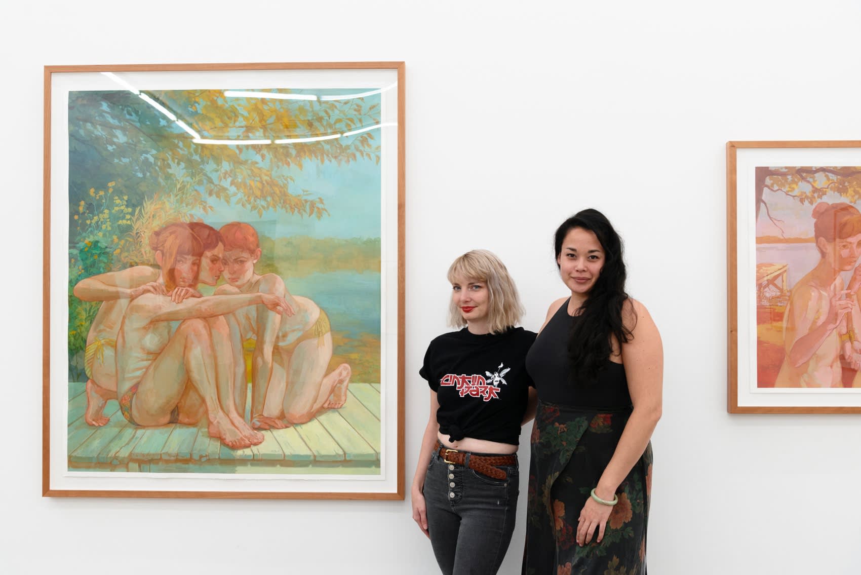Artist Rachel Gregor and LA Director Dasha Matsuura stand together between two of Rachel's framed paintings, where nude girls crouch together, plotting against something slightly out of view. 