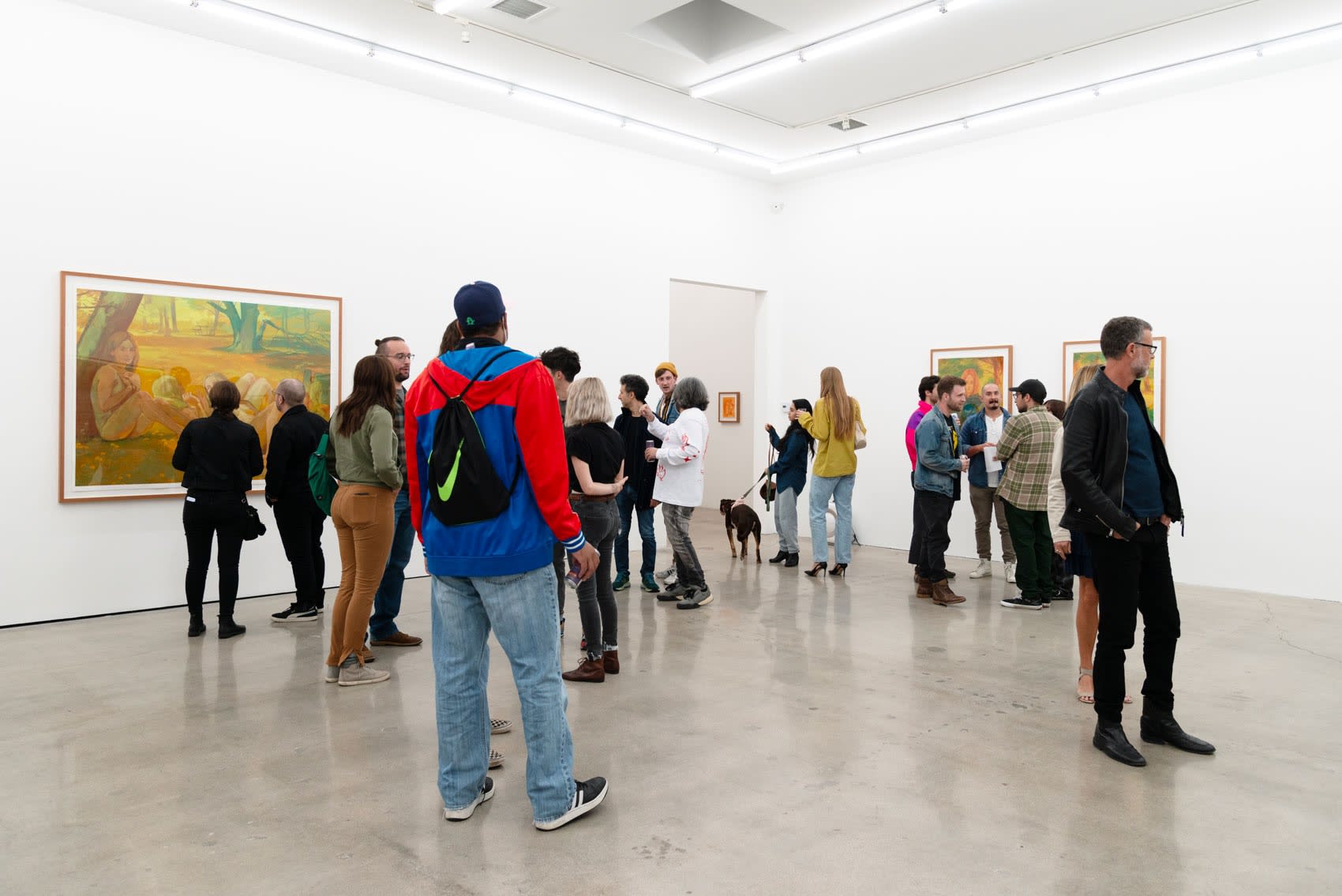 A crowd of about thirty young people gathers inside a white wall art gallery with skylights for the opening reception of Rachel Gregor's painting exhibition, Still Summer.