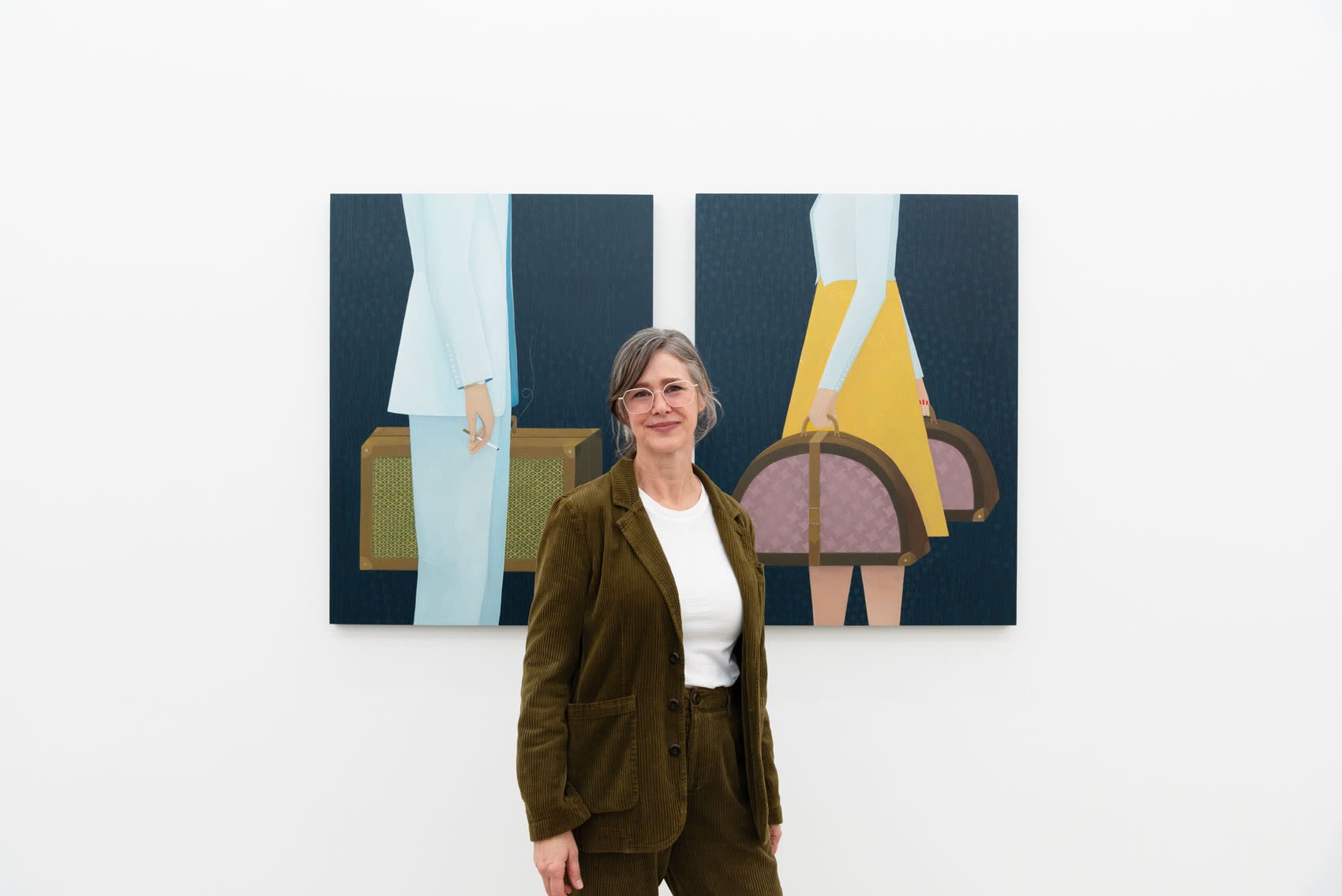 Angela Burson, a white woman with grey hair, stands in front of her paintings of two headless figures with suitcases insdie an art gallery with white walls. 