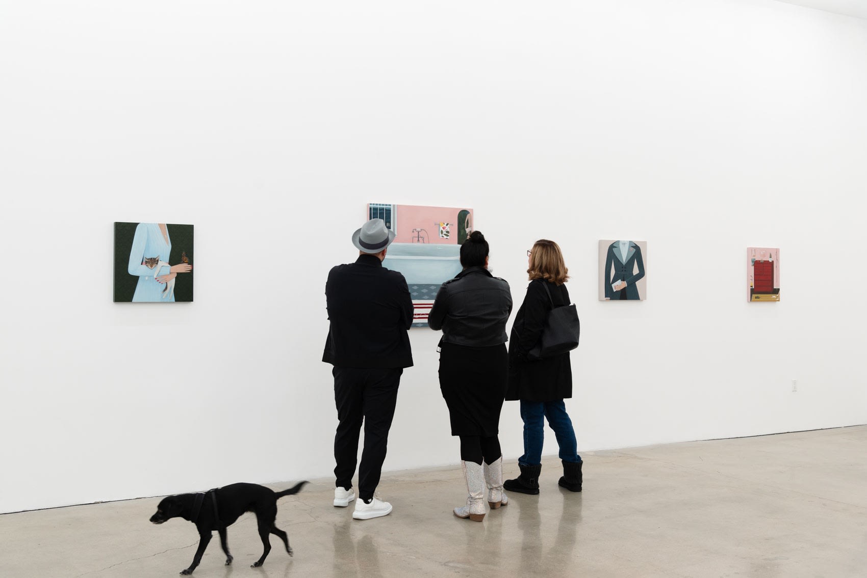 Three people stand up close looking at one of Angela Burson's paintings in an art gallery with white walls, while a small black dog runs away in the bottom left corner. 