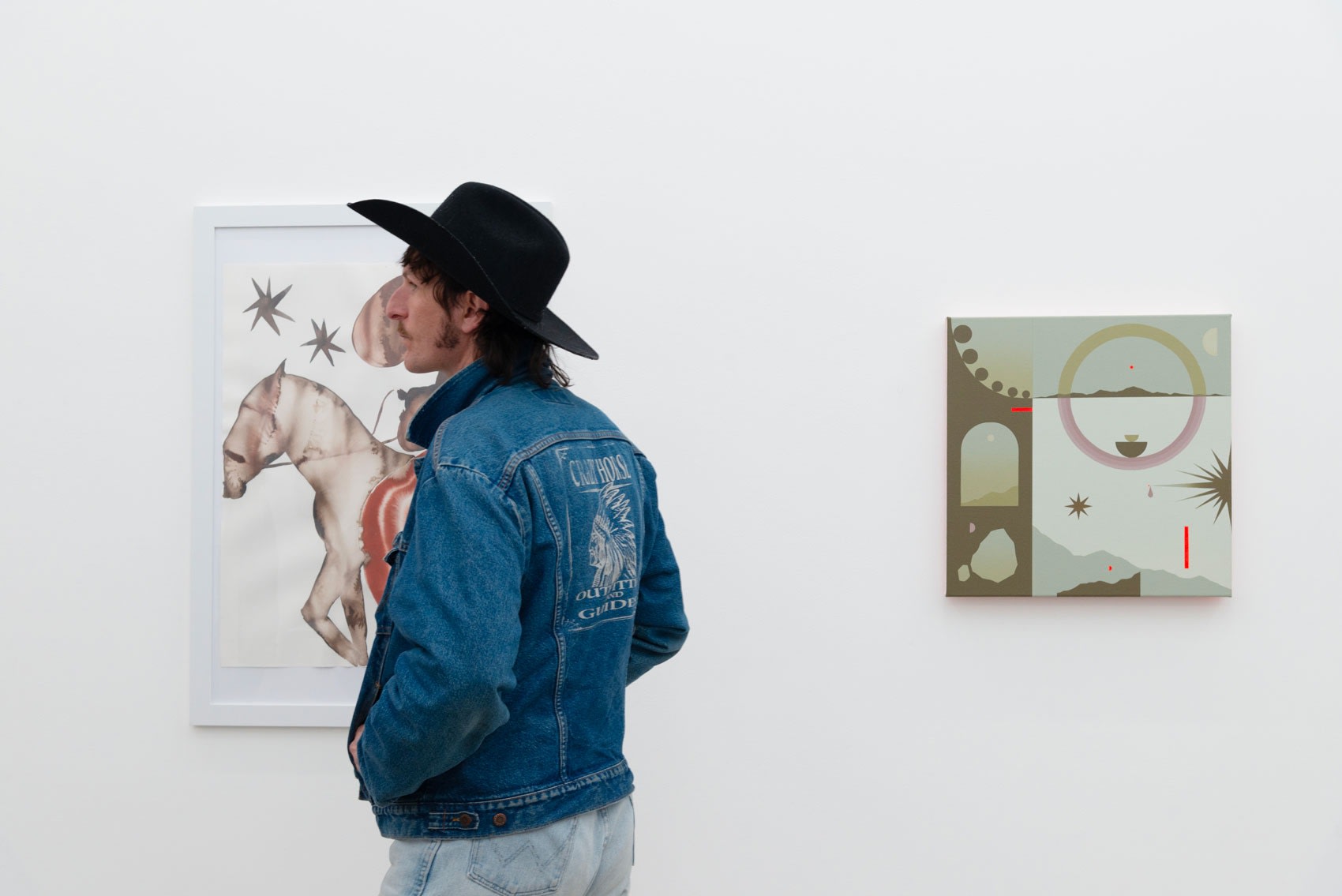 Photo of a man wearing a jean jacket and a black cowboy hat in front of two paintings at Hashimoto Contemporary Los Angeles