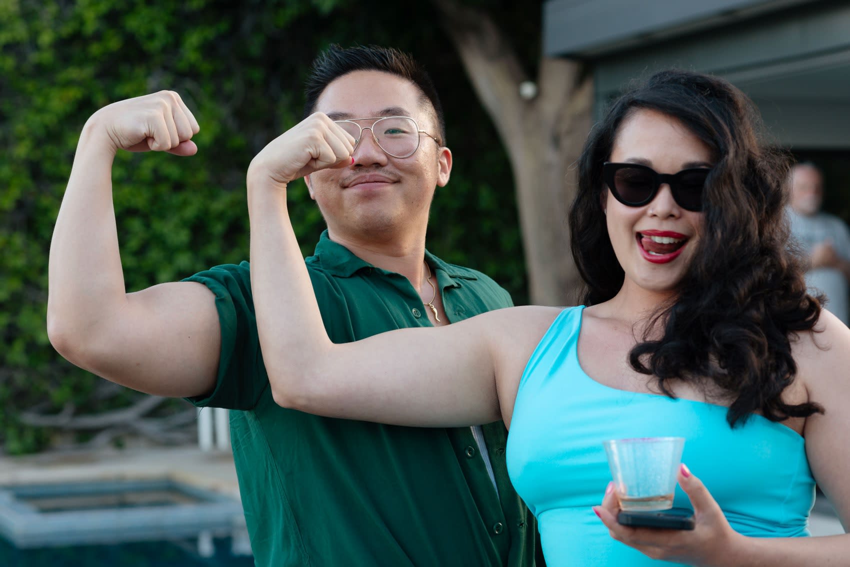 Justin Yoon and Dasha Matsuura flex their biceps in front of an outdoor pool on a summer day