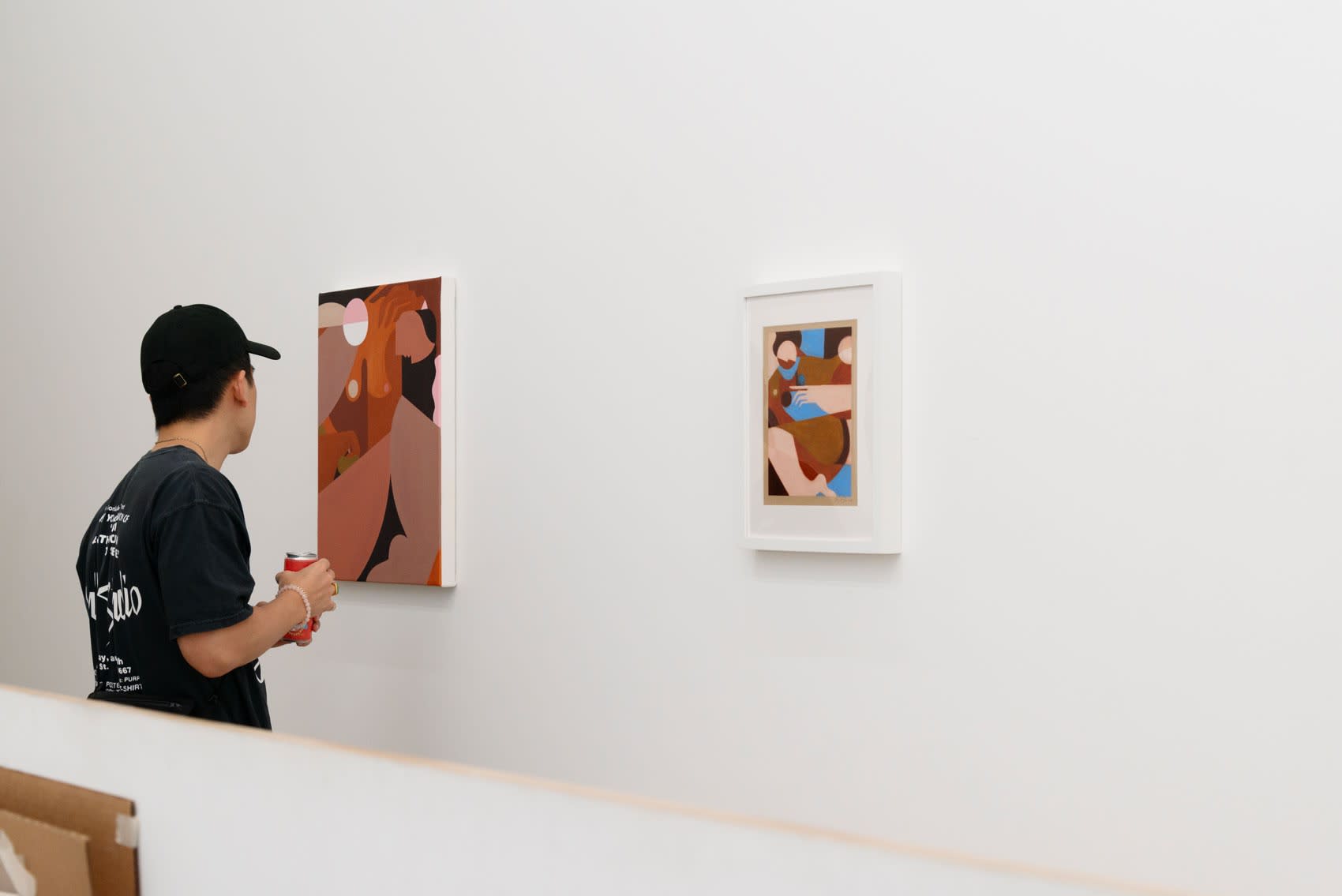 A man in a ball cap and black shirt hold a drink and looks closely at Liz Flores's painting of a woman attempting to scratch her back, rendered in brown, pink, and white neutral tones.
