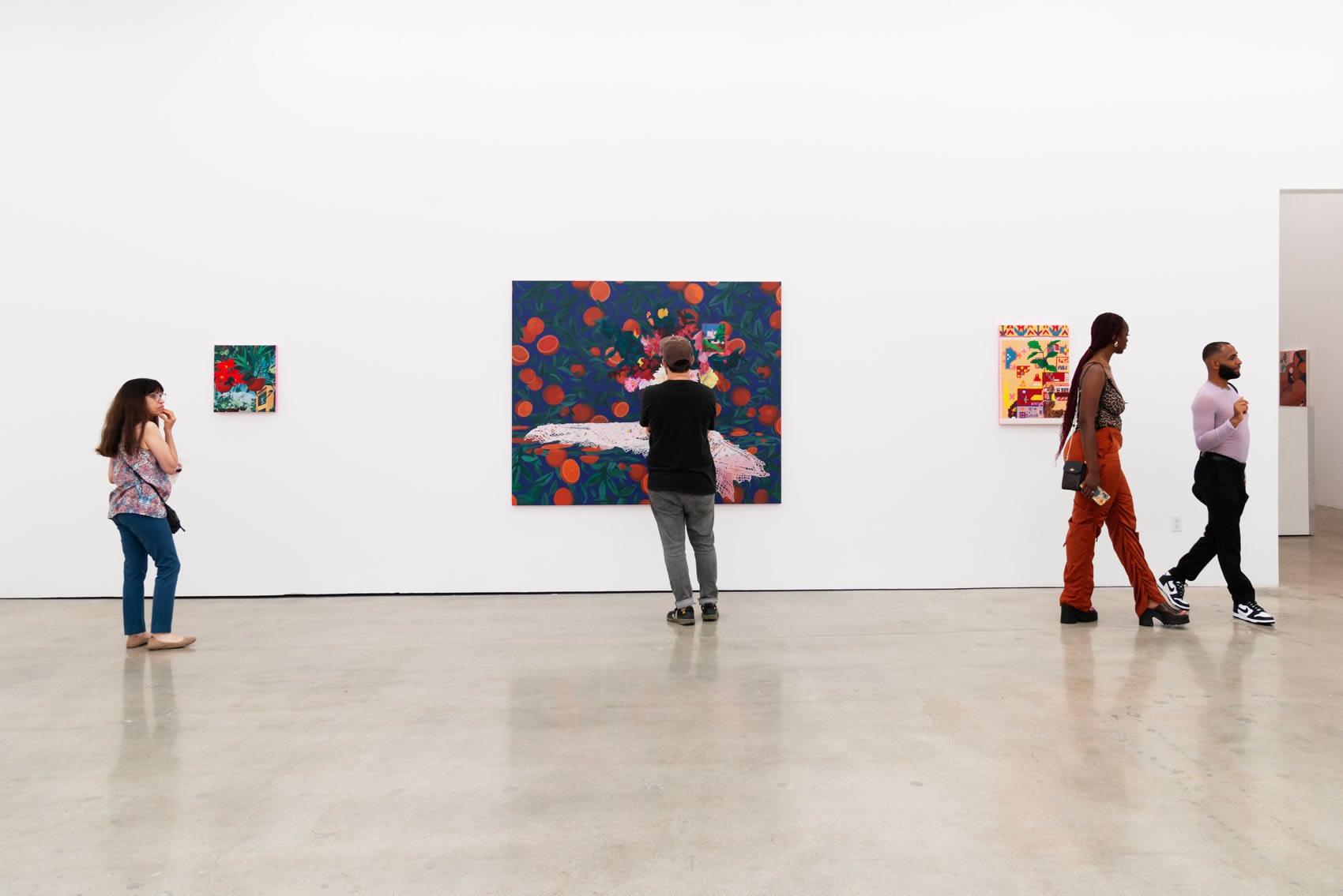 Attendees at the opening of Francisco Díaz Scotto's solo show. Four paintings hang on a white wall, the center one is large, a blue background with oranges and a bouqet, the other three are much smaller and hard to see.