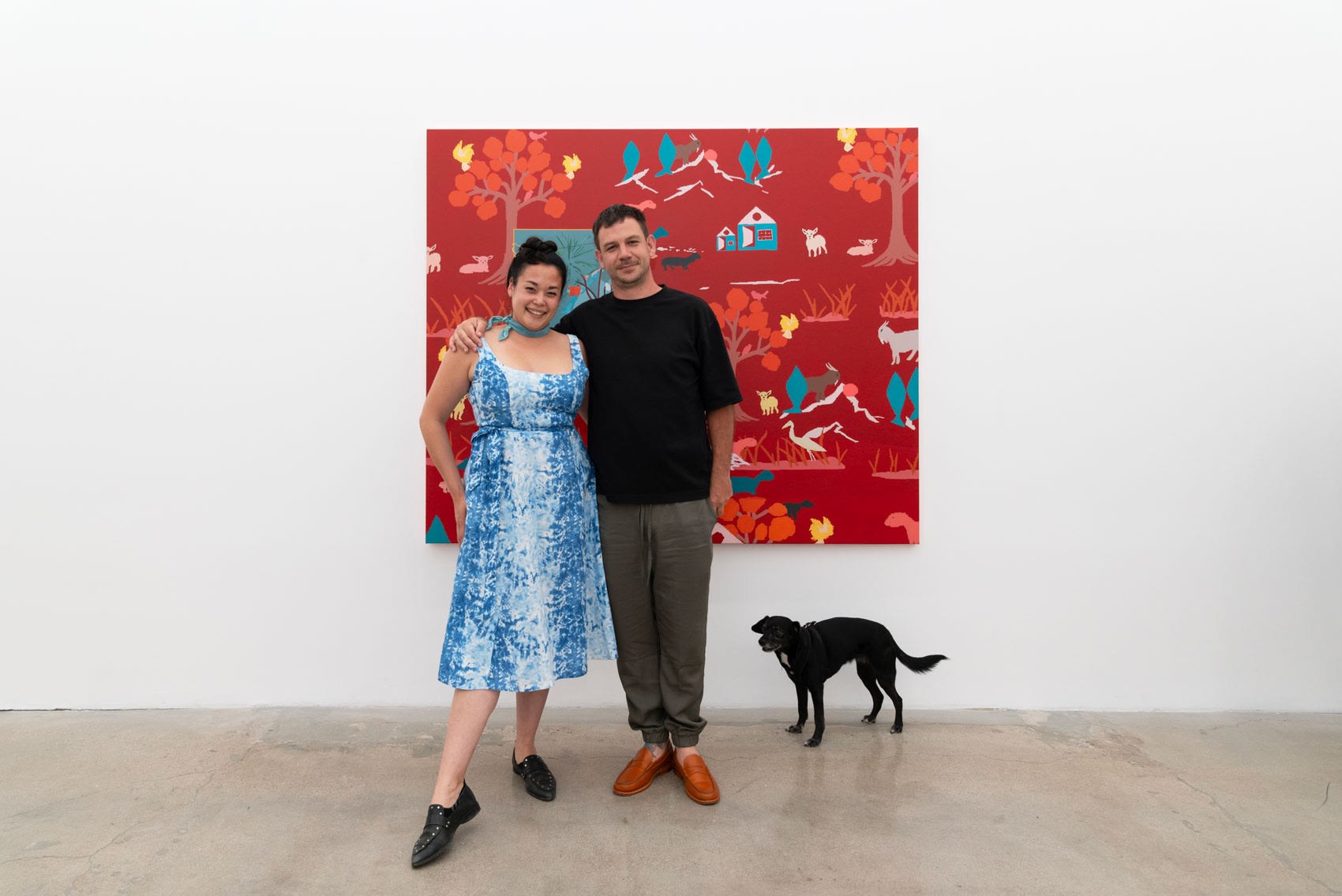 Dasha Matsuura poses in a blue and white dress with Francisco Díaz Scotto, who wears a black weater and white cat eye sunglasses, in front of his large red painting on a white wall. Meera, a small black dog, walks towards them in the bottom right of the frame. 