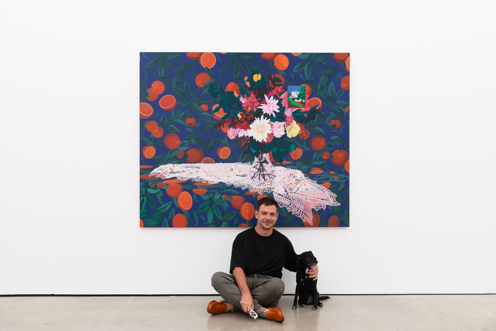 Artist Francisco Diaz Scotto with a small black dog, Meera, sitting on the ground in front of one of his large scale paintings, which is mostly blue and orange, with a bright bouquet of flowers on a white lace table cloth in the middle of the composition.