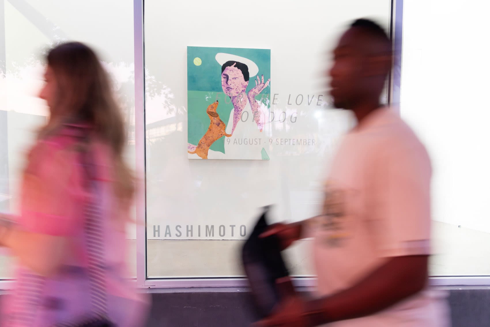 Erin Armstrong's painting of a woman in a hot with a wienr dog is seen through a window as two blurry people walk past
