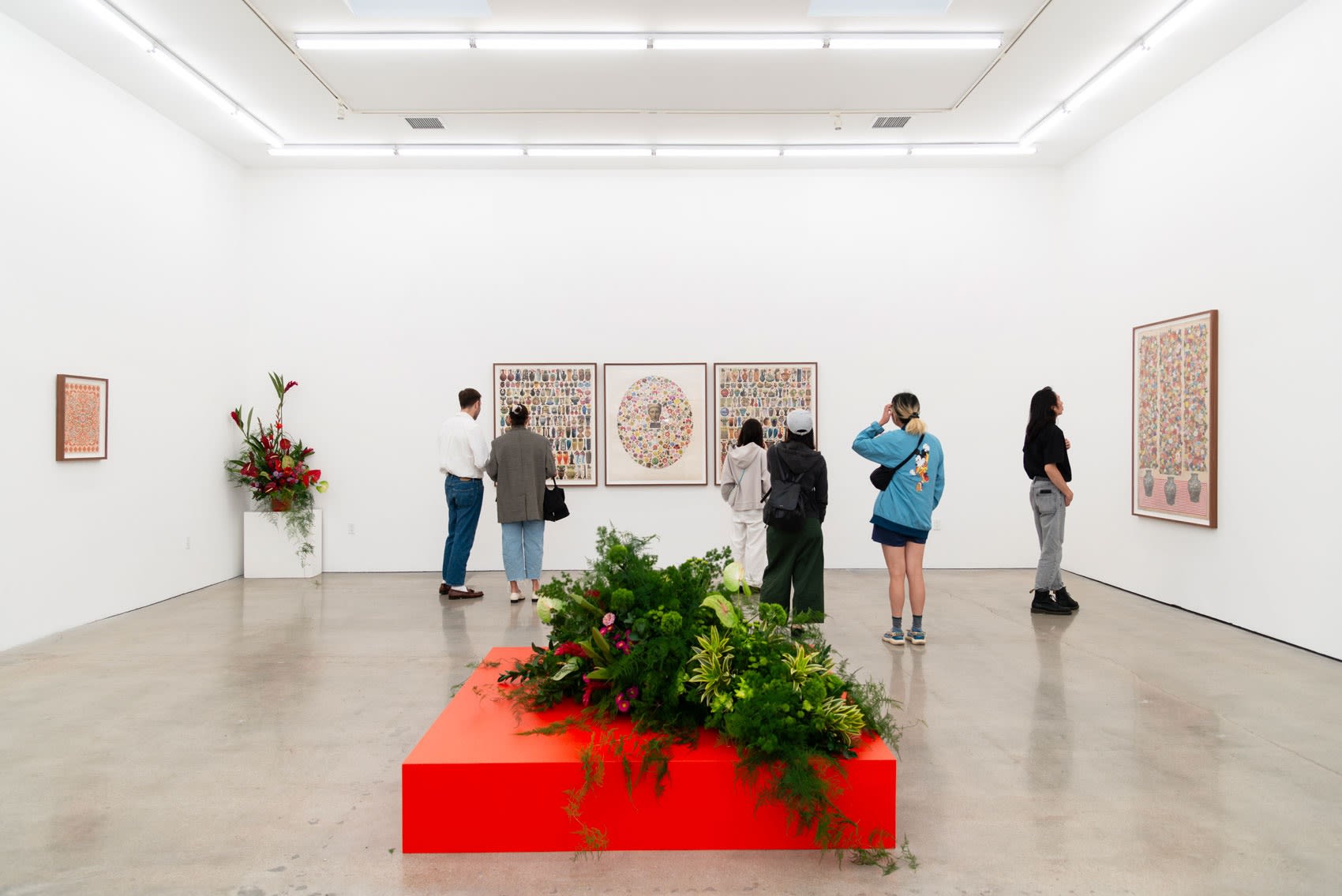 Attendees of matthew Craven's solo show look at the large collage and drawn works on paper in a white cube gallery space with concrete floors. A floral installation on a brighr red pedestal is in the middle of the room 