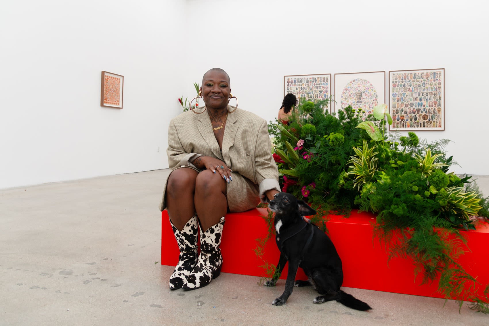 Flora Tosha Stimage sits on a red box next to her flower installation petting a small black dog in the gallery space
