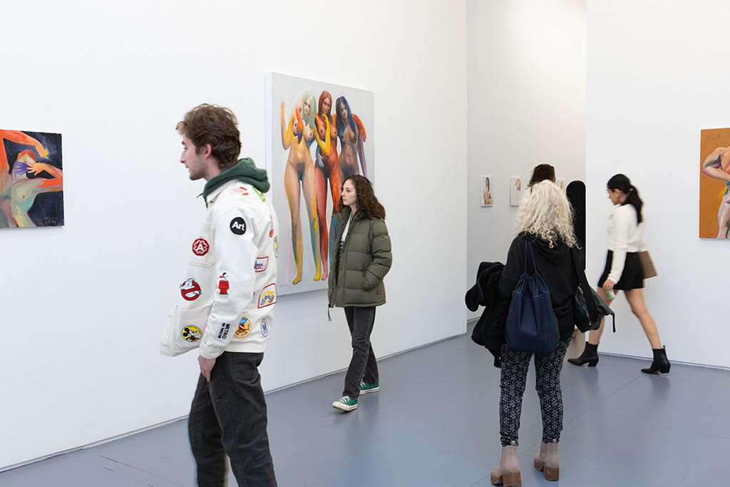 A crowd of people gather inside Erik Jones's solo exhibition Muse inside a New York City art gallery with white walls and florescent lighting. 