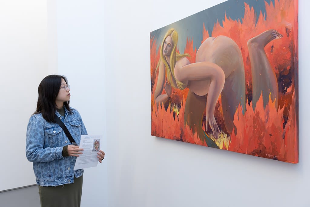 A woman looks at Erik Jones's painting of a white woman with blonde hair smiling inside a hellish fire. 