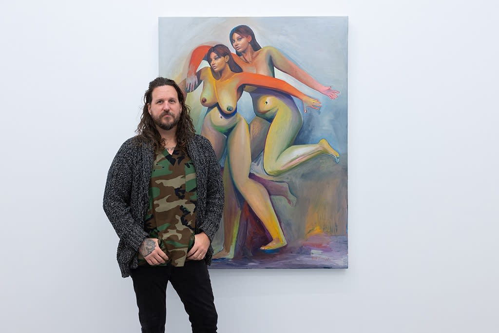 Erik Jones, a white man with long hair wearing a camo print shirt, stands in front of his painting with two colorful nude women. He stands in a white wall art gallery with white concrete floors. 