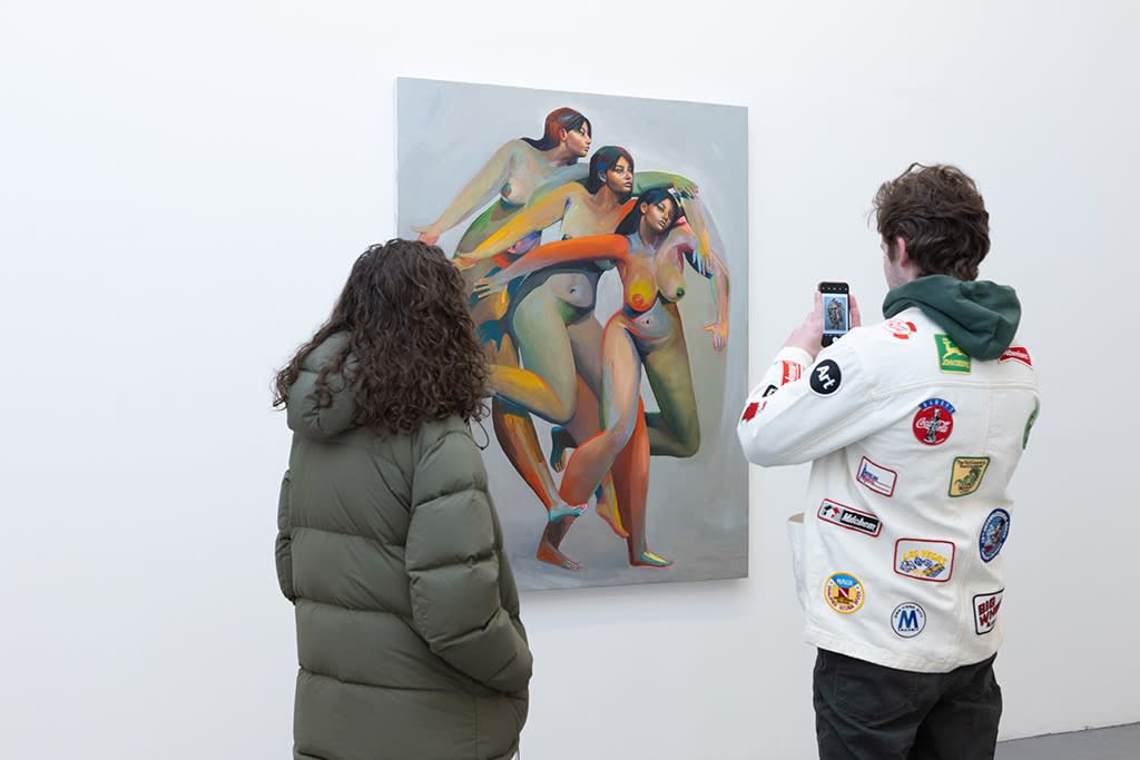 Two people stand and take pictures of a painting by Erik Jones of three nude colorful women descending the painting's frame. 