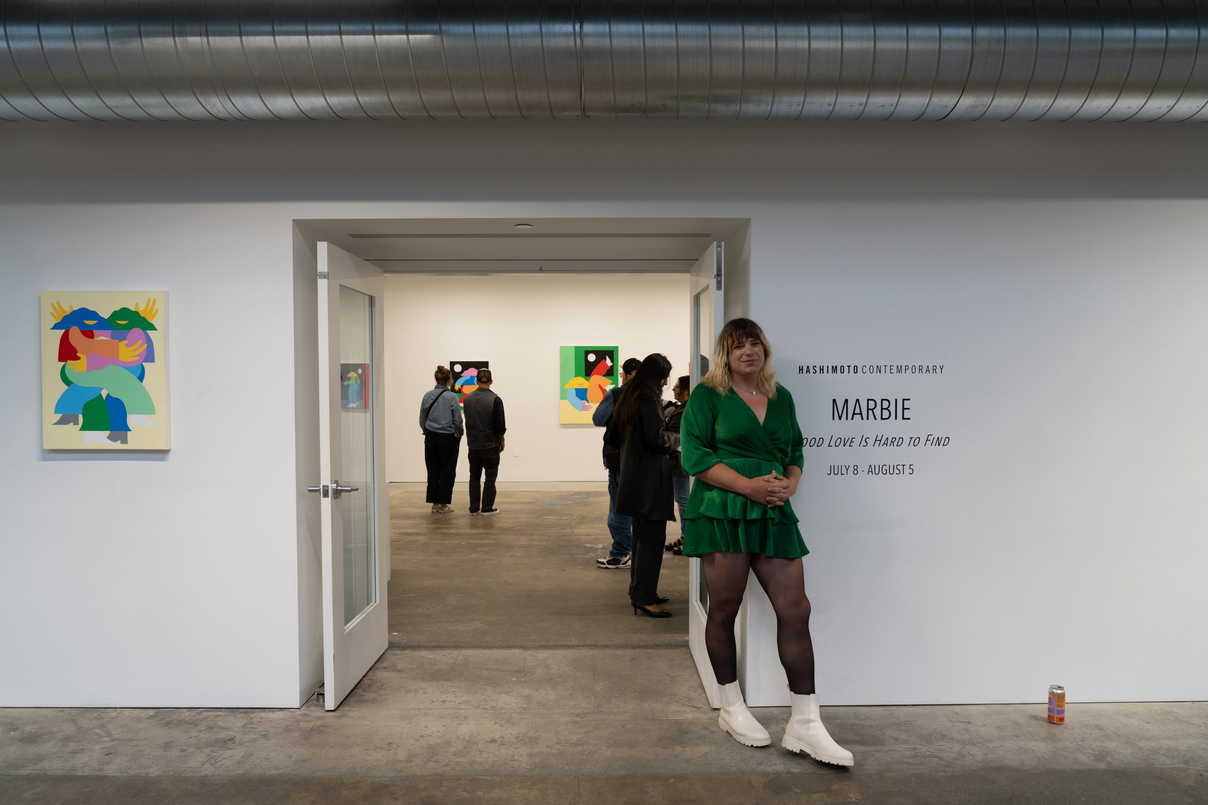 Marbie stands in front of the doorway to her painting show, wearing a green velvet skater dress, black tights, and white boots