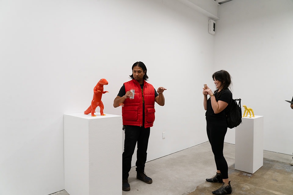 A woman in a black shirt takes a photo of a man in a red vest who is imitating a red dinosaur standing on its back legs, showing off its yellow nails.