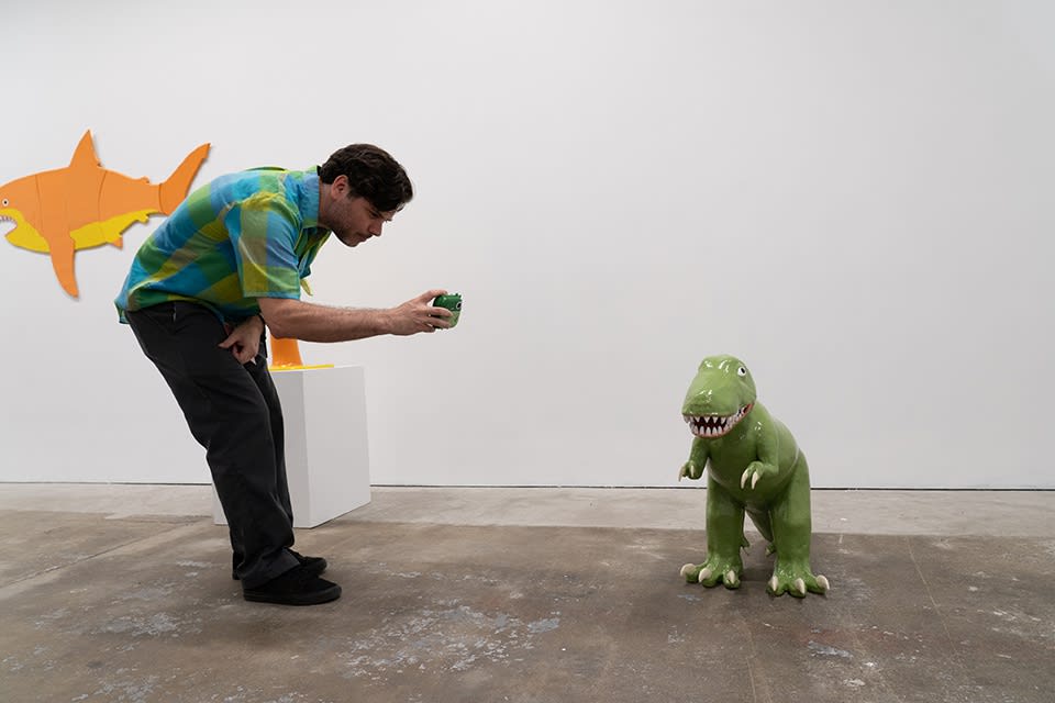 A men stands in a white cube gallery, bending over to take a picture of a ceramic stegasaurus on his phone