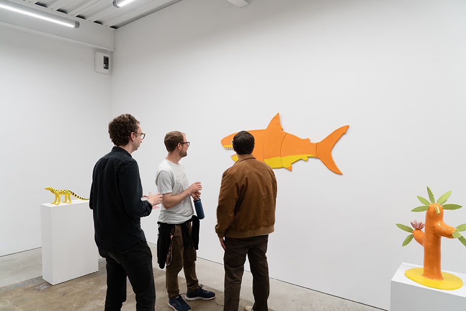 Three men stand and admire an orange megalodon ceramic wall hanging against a white wall and concrete floor. 