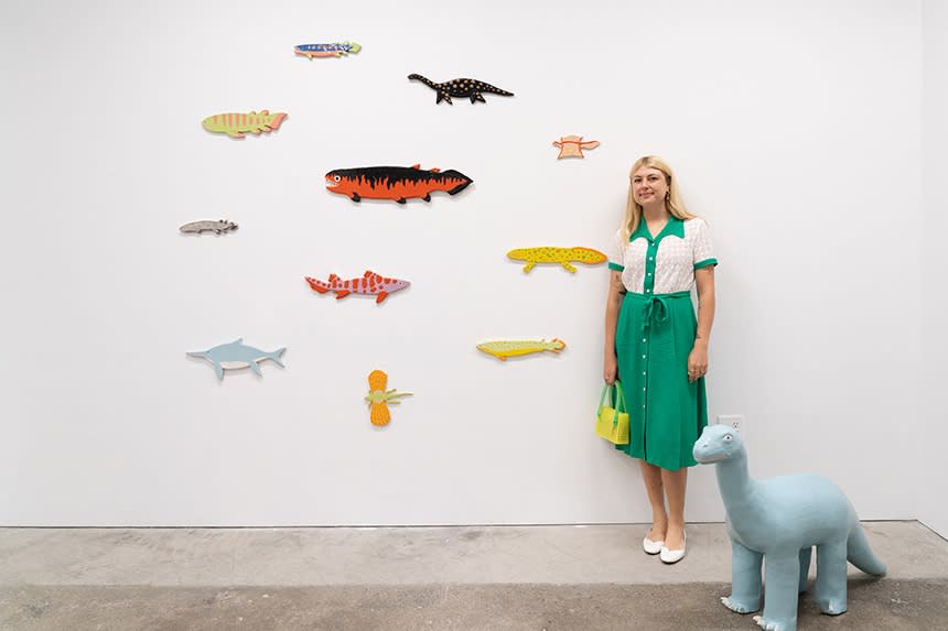 artist Lorien Stern in a white and green dress stands with her artworks, which are ceramic dinosaurs and wall hangings, at the opening of her solo show Old Friends.