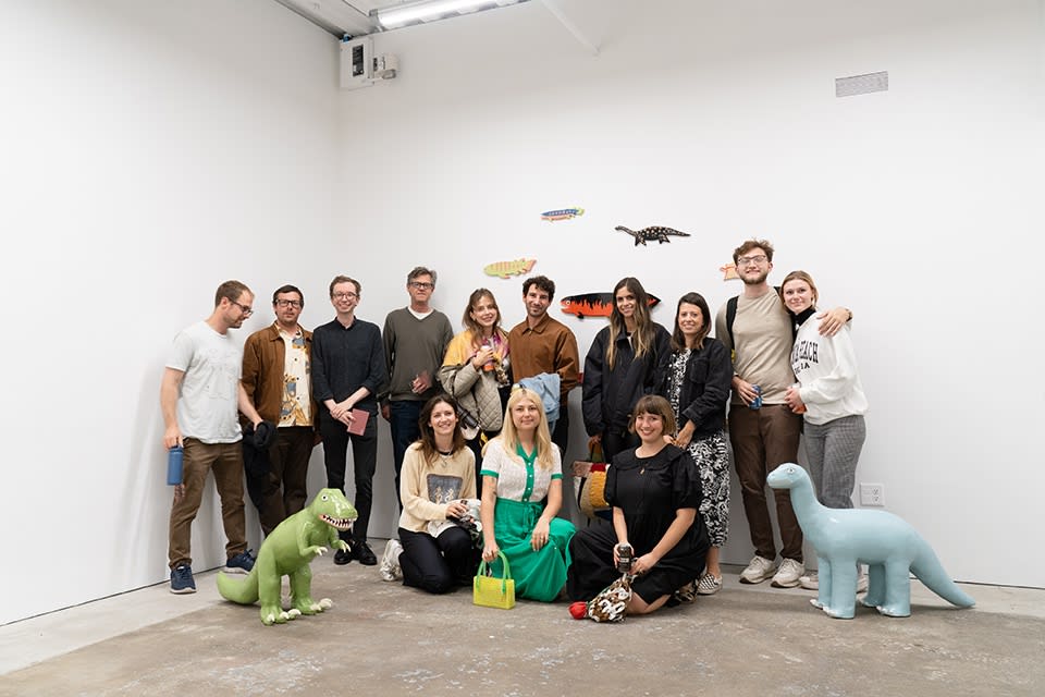 A group of Lorien Stern's friends and family gather for a group picture in a white cube gallery, with wall hangings of small fish behind them