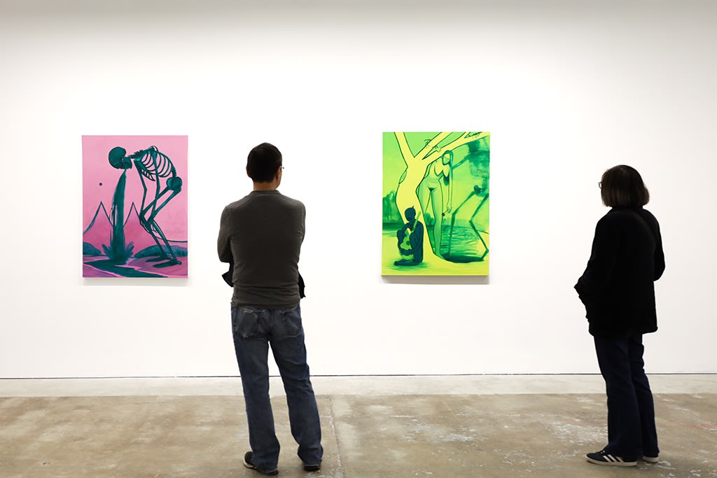 Two people stand in a white wall gallery with concrete floors looking at two paintings: one pink with a dark green skeletong throwing up and another bright green and yellow of a nude woman approaching a crying clown from around a tree while a dark skeleton looms over the scene. 