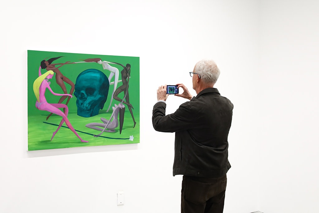An older man stands in front of a green paintings with colorful nude women dancing in front of a dark green skull, taking a horizontal picture with his phone. 