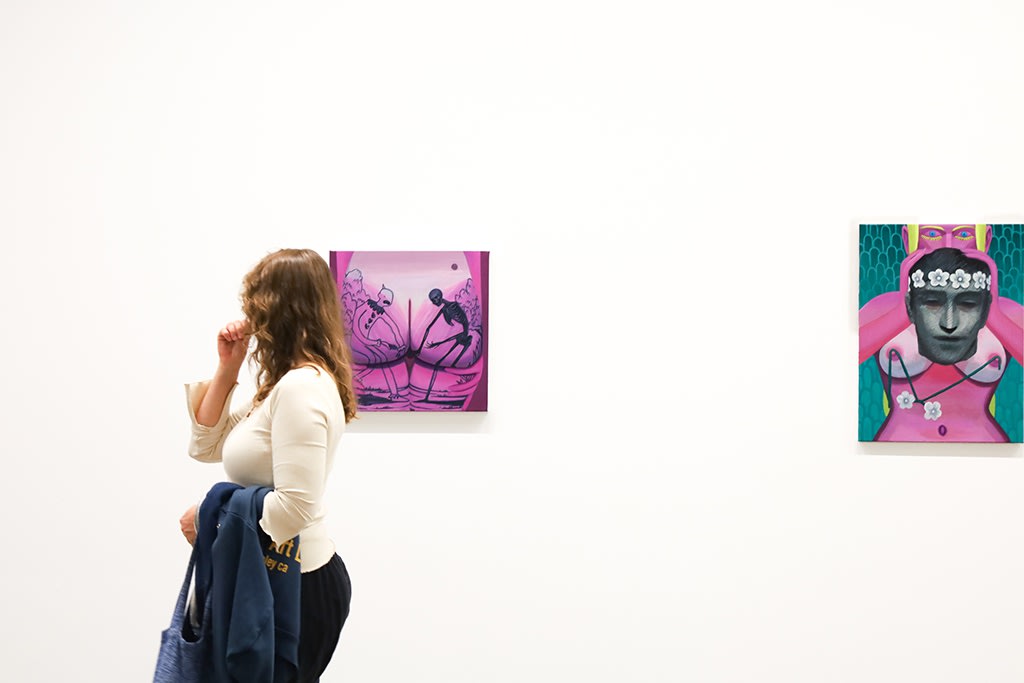 A woman holds her coat and walks past two paintings on a white wall: one is a pink paintings of a buttocks where a clown on one cheek and a dark skeleton on the other grab a cheek to enter, the other is a painting of a women facing the viewer, holding a dark grey man's head in front of her chest, her yellow lashes framing her wide open blue eyes on pink body.
