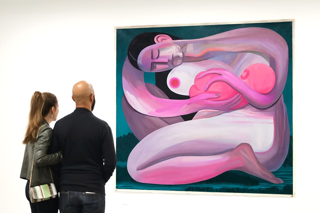 A man and woman stand close to each other in a white wall art gallery looking at a huge painting of a pink woman with black hair cradling her baby