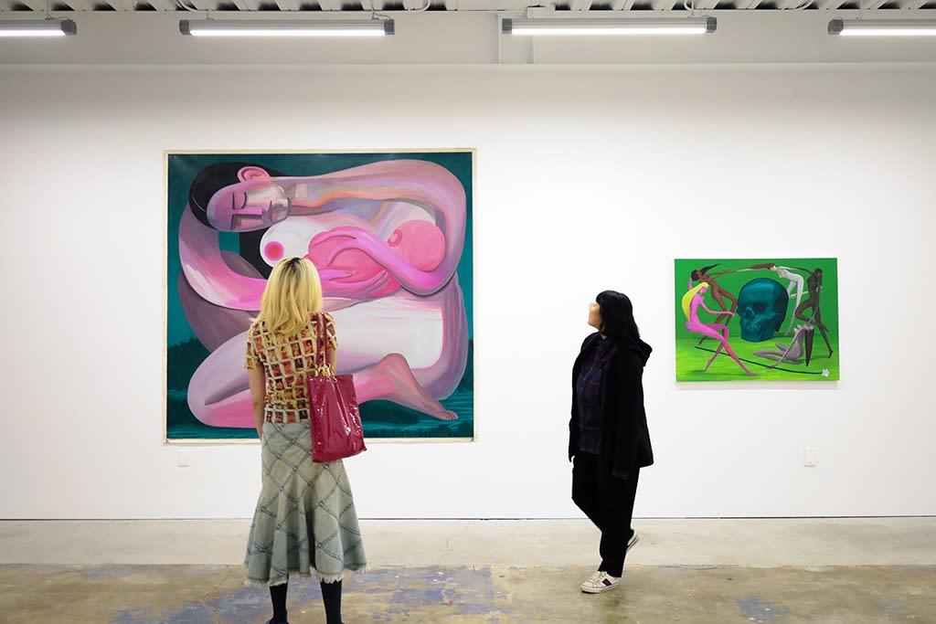 Two people stand in a white wall gallery with concrete floors looking at two small, brightly colored paintings obscured by how far away they appear.