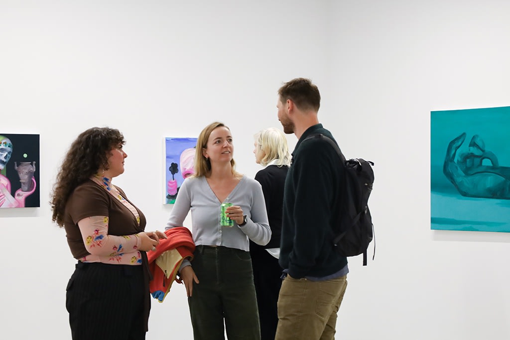 A group of young people stand in a white wall gallery with concrete floors talking and laughing. In the background are two paintings: one green with nude figures dancing in a cirlce, and another bright pink with a dark green skeleton throwing up. 