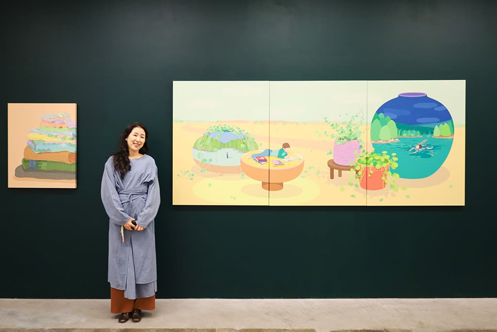Danym Kwon, a Korean woman in a blue dress, stands next to her large triptych painting of a boy reading on a surreally large bowl in the dessert. The painting hangs on a dark green wall in an art gallery. 