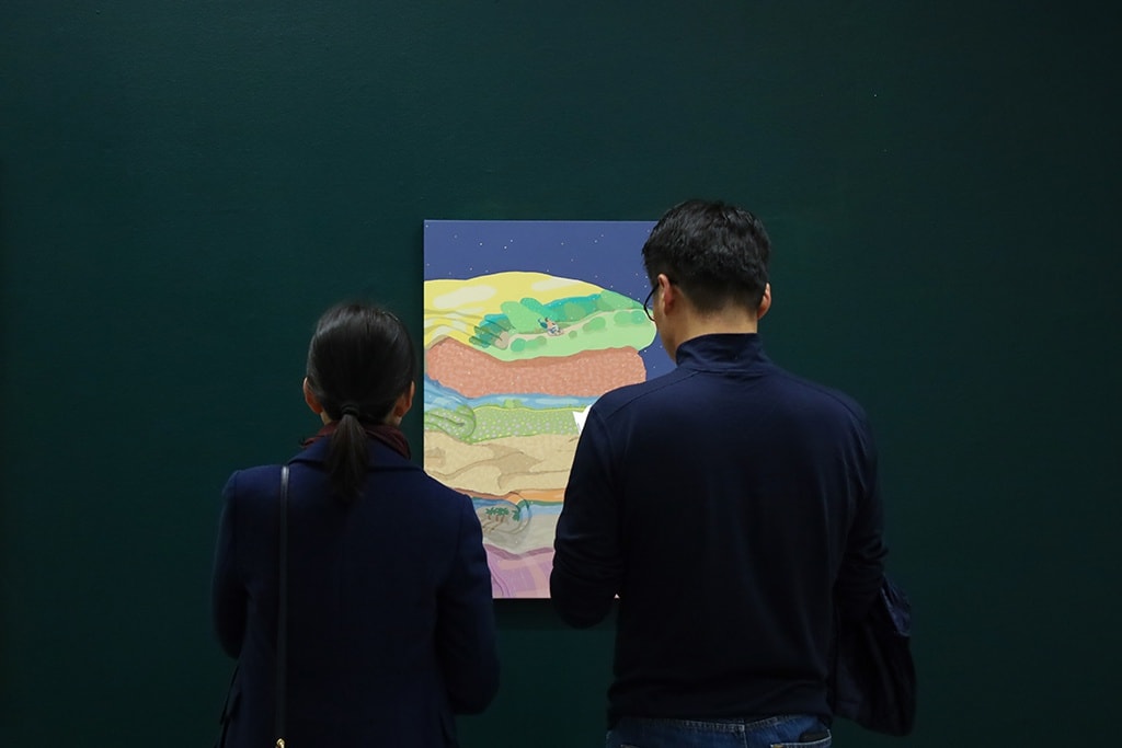 two people stand and look at Danym Kwon's painting of pastel colored folded laundry on a dark blue baackground. The painting hangs on a forest green wall in an art gallery. 