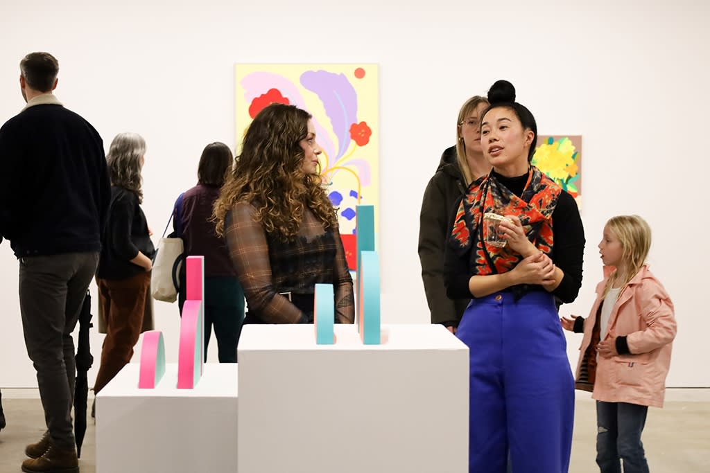 Terri Chiao of CHIAOZZA speaks to Rachel Strum in front of her colorful resin sculptures on white plinths at the opening of 