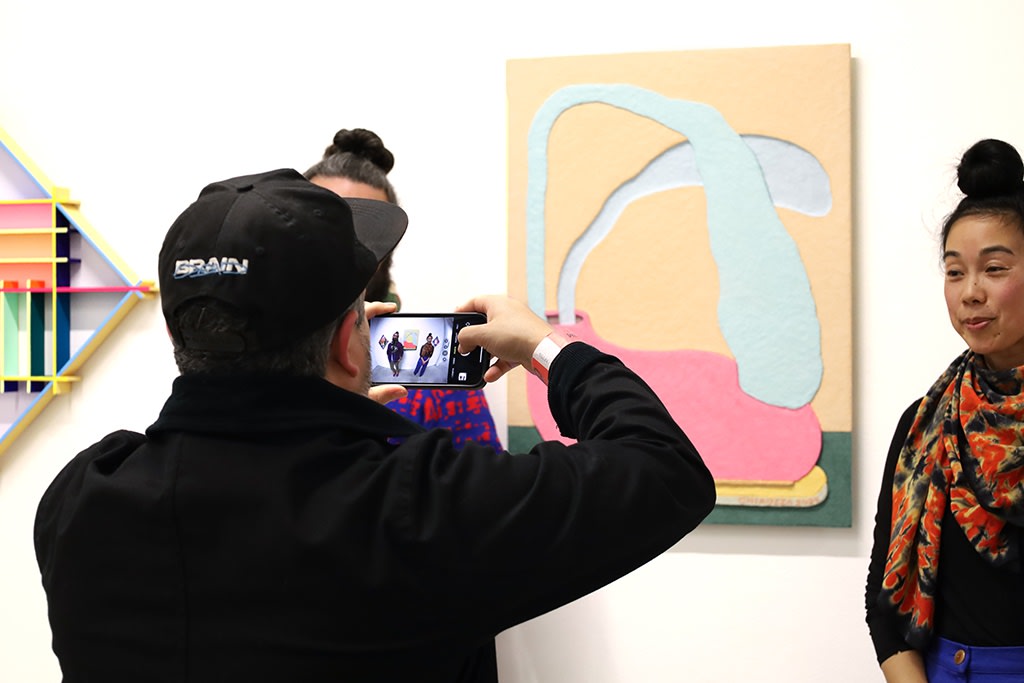 An attendee takes a fish-eye lens photo of the artists with their colorful works inside a white wall gallery with fluorescent lighting. 